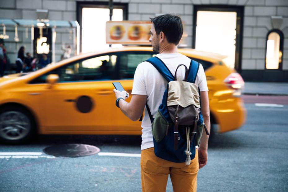A man stands on a city street, looking at his phone, a yellow cab is behind him. 